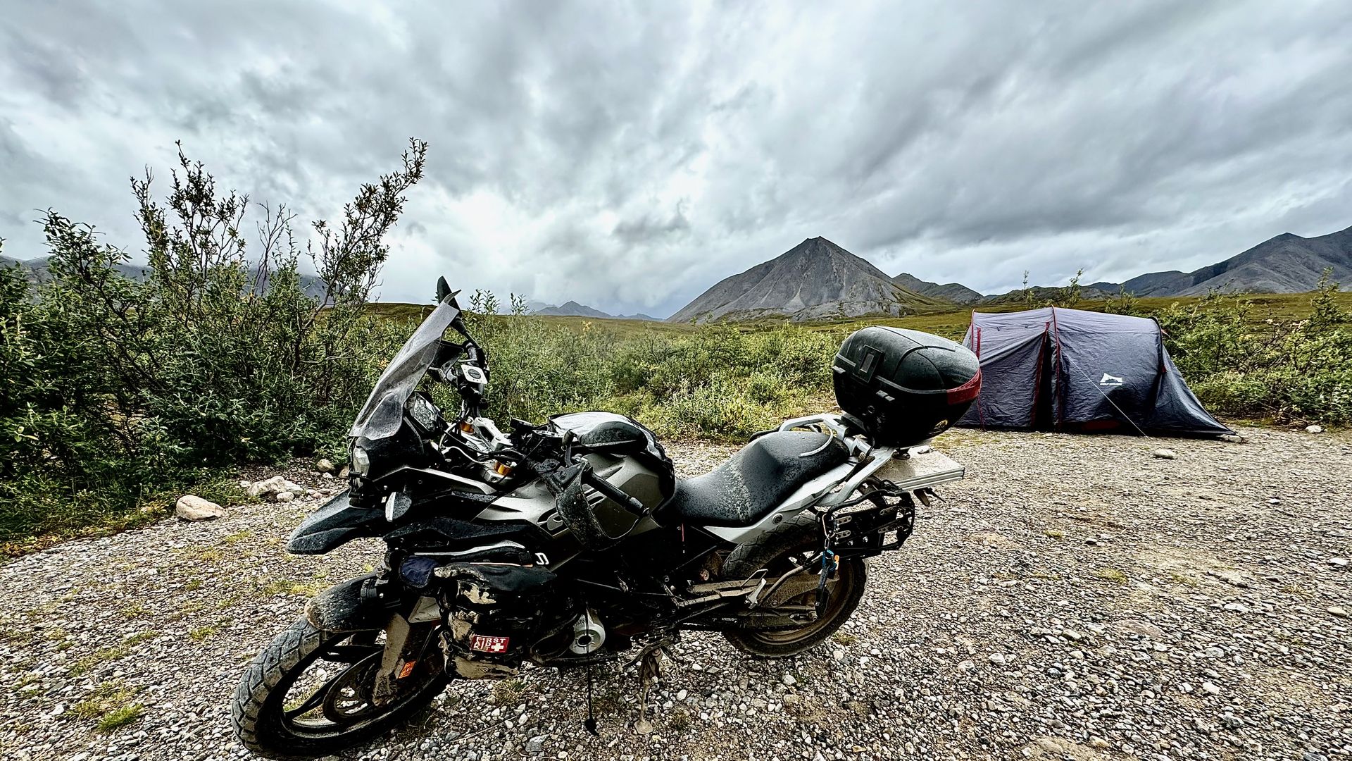 A loaded BMW G310GS parked on a gravel pullout in the Alaskan tundra, a small tent pitched behind it beneath a low storm sky.